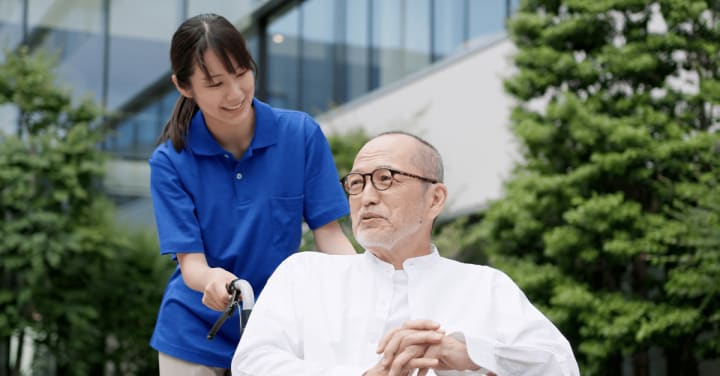 Grateful Hearts: A Year Of Gentle Dementia Care 1 A Smiling Caregiver In A Blue Polo Shirt Walks Outdoors With An Older Man Sitting In A Wheelchair.