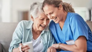 A caregiver in blue scrubs and an older woman share a joyful laugh together indoors.