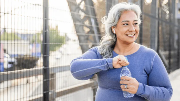 A smiling older woman with gray hair opens a bottle of water while wearing athletic clothing outdoors.