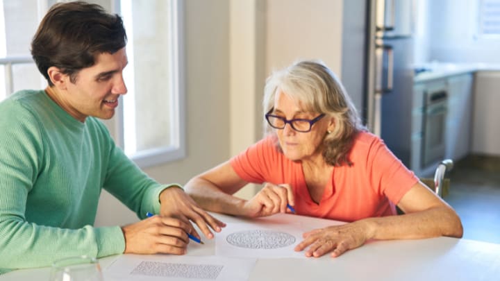 &Quot;Mom Keeps Forgetting&Quot;: How Our Dallas Memory Care Community Helps Families Cope 1 A Young Man In A Green Sweater Sits At A Table And Helps An Older Woman Complete A Paper Maze Puzzle.