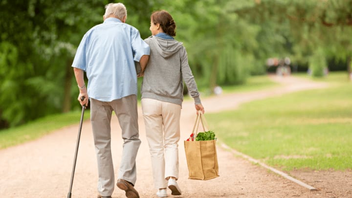 How A Memory Care Community In Dallas, Tx Enhances Support Systems 1 Rear View Of A Woman Carrying Groceries While Walking Arm-In-Arm With An Elderly Man Using A Cane In A Park.