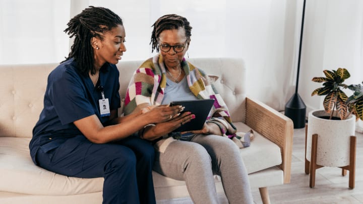 Providing Exceptional Care For Memory Loss Residents In Dallas: Why It Matters 1 A Caregiver In Dark Blue Scrubs Sits On A Couch And Helps An Older Woman Use A Digital Tablet.