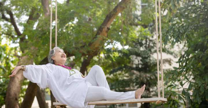 Memory Care Sensory Sanctuary In Turtle Creek 1 An Older Woman In White Clothing Smiles Joyfully While Leaning Back On A Large Wooden Rope Swing Surrounded By Green Trees.
