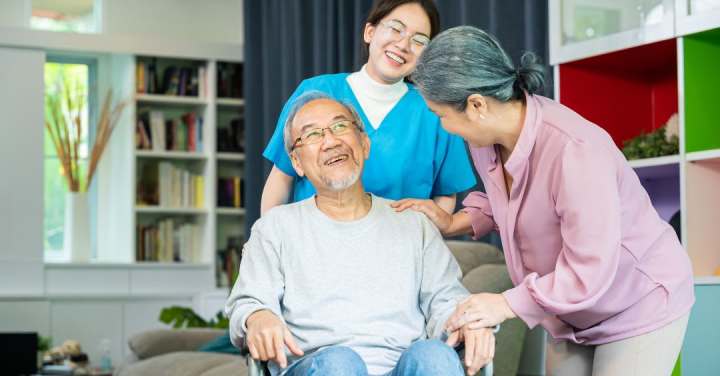 Specialized Caregivers Are The Gold Standard For Memory Care 1 A Smiling Caregiver In Blue Scrubs And An Older Woman Support An Elderly Man Seated In A Chair As They All Share A Warm, Joyful Moment.