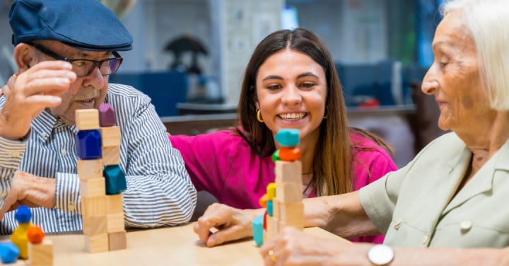 Dementia Care: Boost Senior Skill-Building Now 1 A Smiling Caregiver Assists An Older Man And Woman Playing A Tabletop Block Stacking Game.