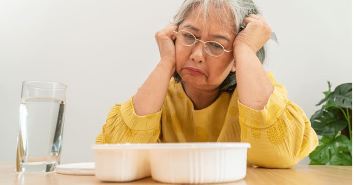 Appetite Loss In Memory Care: Warning Signs &Amp; Solutions 1 An Older Woman Looks Down With A Sad, Reluctant Expression At A Tray Of Food On The Table In Front Of Her.