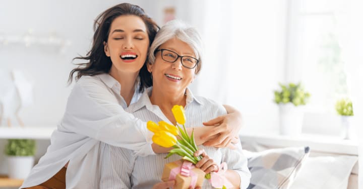 Lasting Love: Mother'S Day In Memory Care 1 A Younger Woman Joyfully Hugs An Older Woman From Behind As They Smile Together, With The Older Woman Holding A Small Gift And Yellow Tulips.